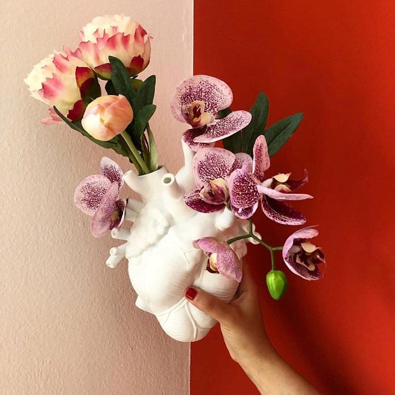 Hand holding a white vase with pink flowers against a red and beige wall.
