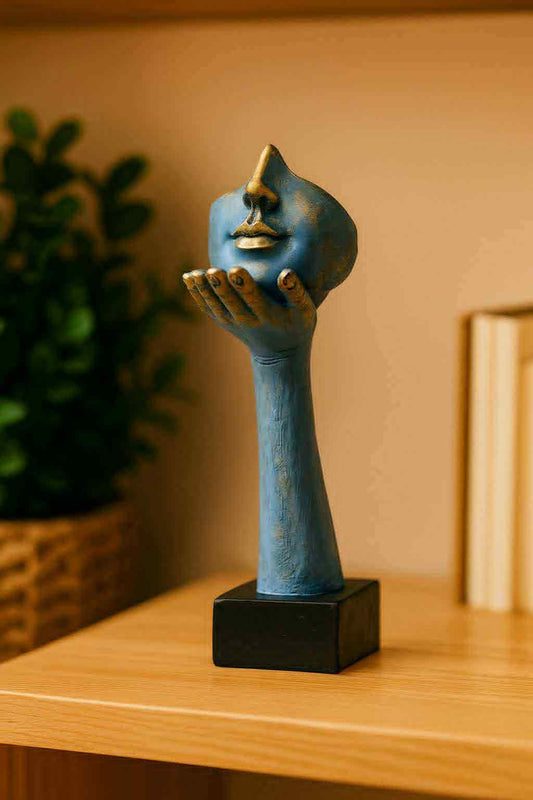 Blue sculpture of a hand holding a face on a wooden surface with a plant and books in the background.