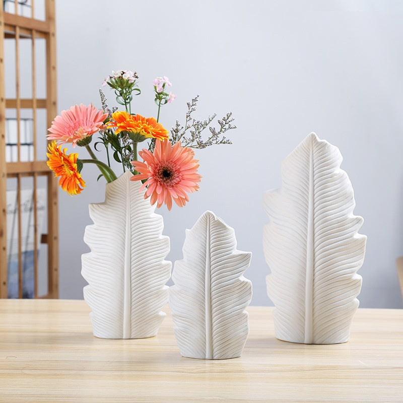 Set of three white leaf-shaped vases with flowers on a wooden surface.