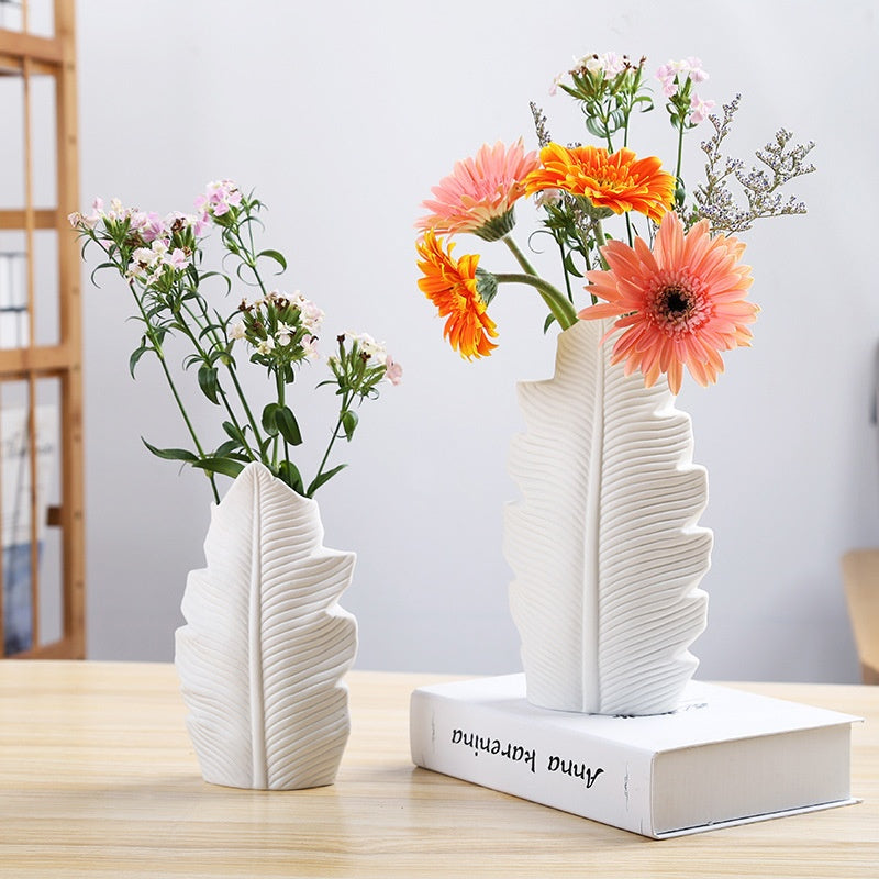 Two white leaf-shaped vases with flowers on a wooden table.