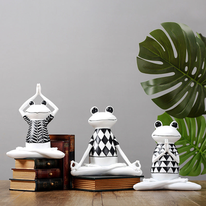 Three frog statues in various yoga poses on books with a plant in the background