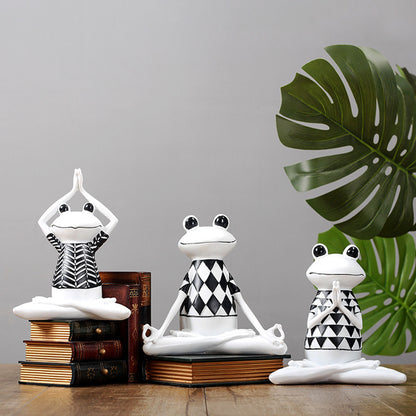 Three frog statues in various yoga poses on books with a plant in the background